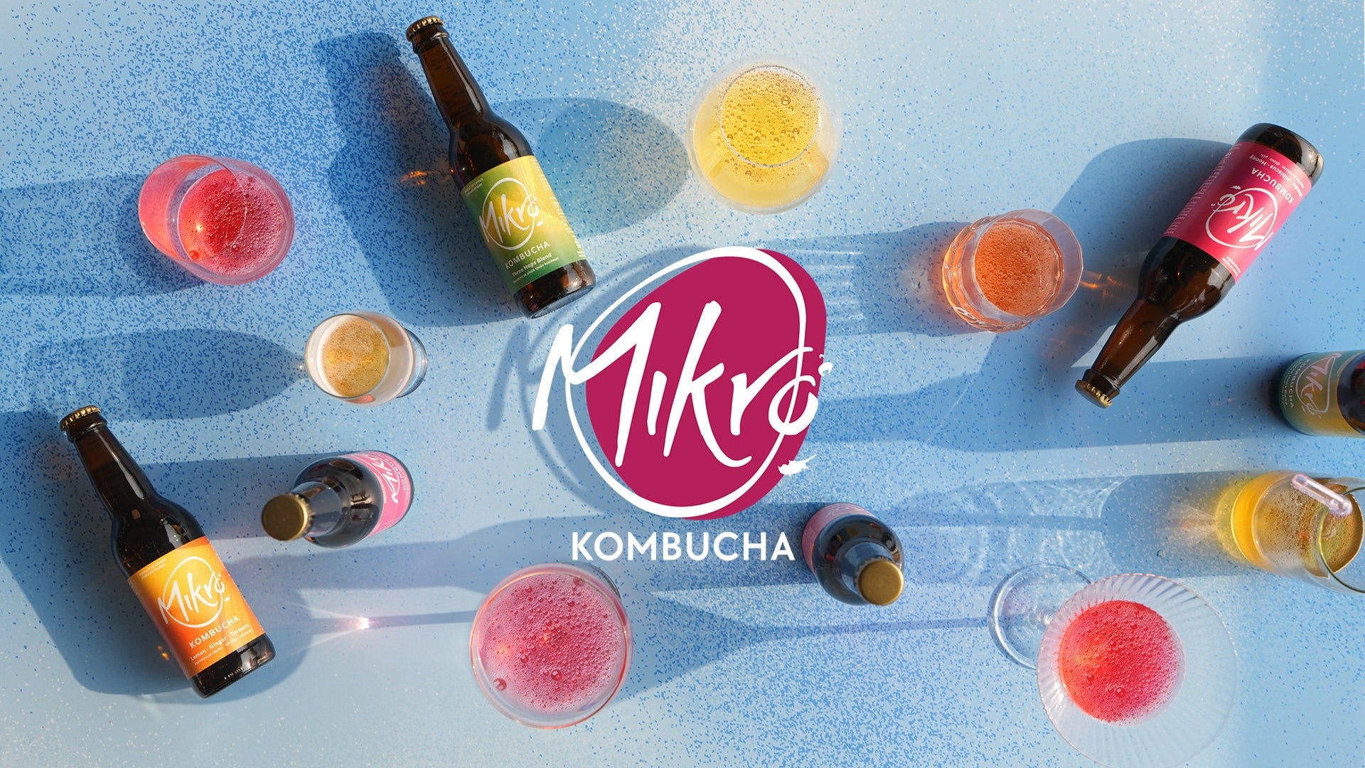 Mikro Kombucha bottles and glasses on a blue surface with the Mikro Kombucha logo.