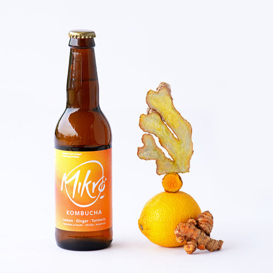 Mikro Lemon Ginger Turmeric kombucha bottle with bright yellow label next to a lemon, ginger and turmeric pieces on a white background.