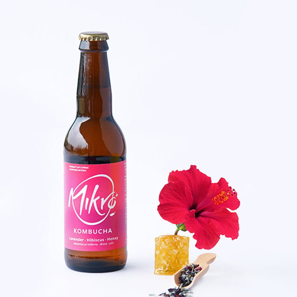 Mikro Lavender Hibiscus Honey Kombucha bottle with bright pink and purple label next to lavender buds, hibiscus flower and honey comb piece on a white background