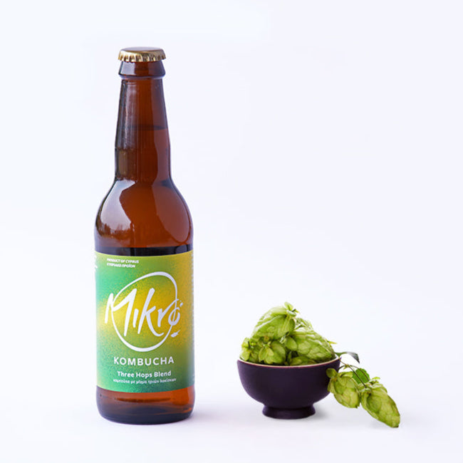 Mikro Three Hops blend kombucha bottle with bright green and yellow label next to a bowl of hops buds on a white background