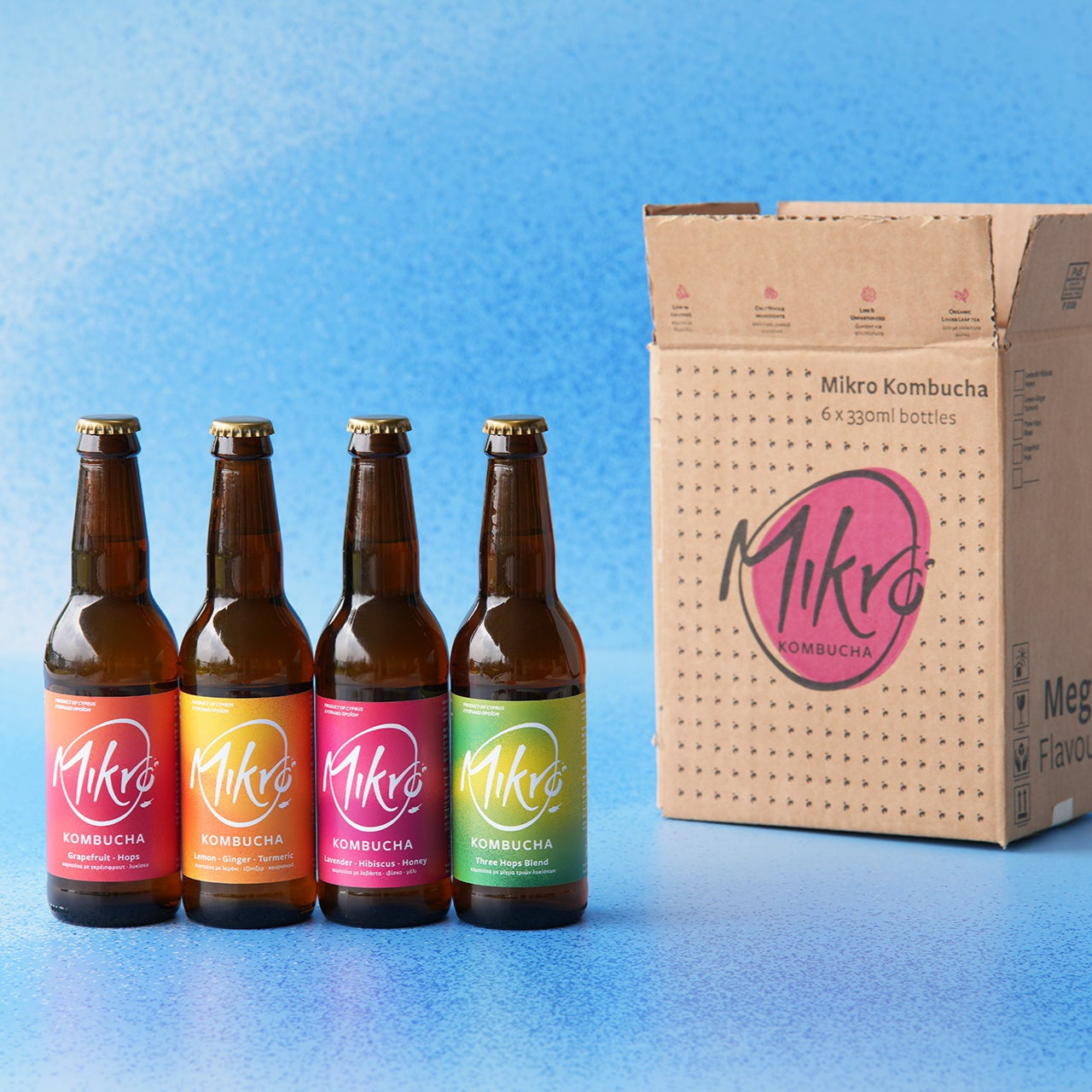 Four bottles of different flavours of Mikro Kombucha with colorful labels next to a cardboard box on a blue gradient background.