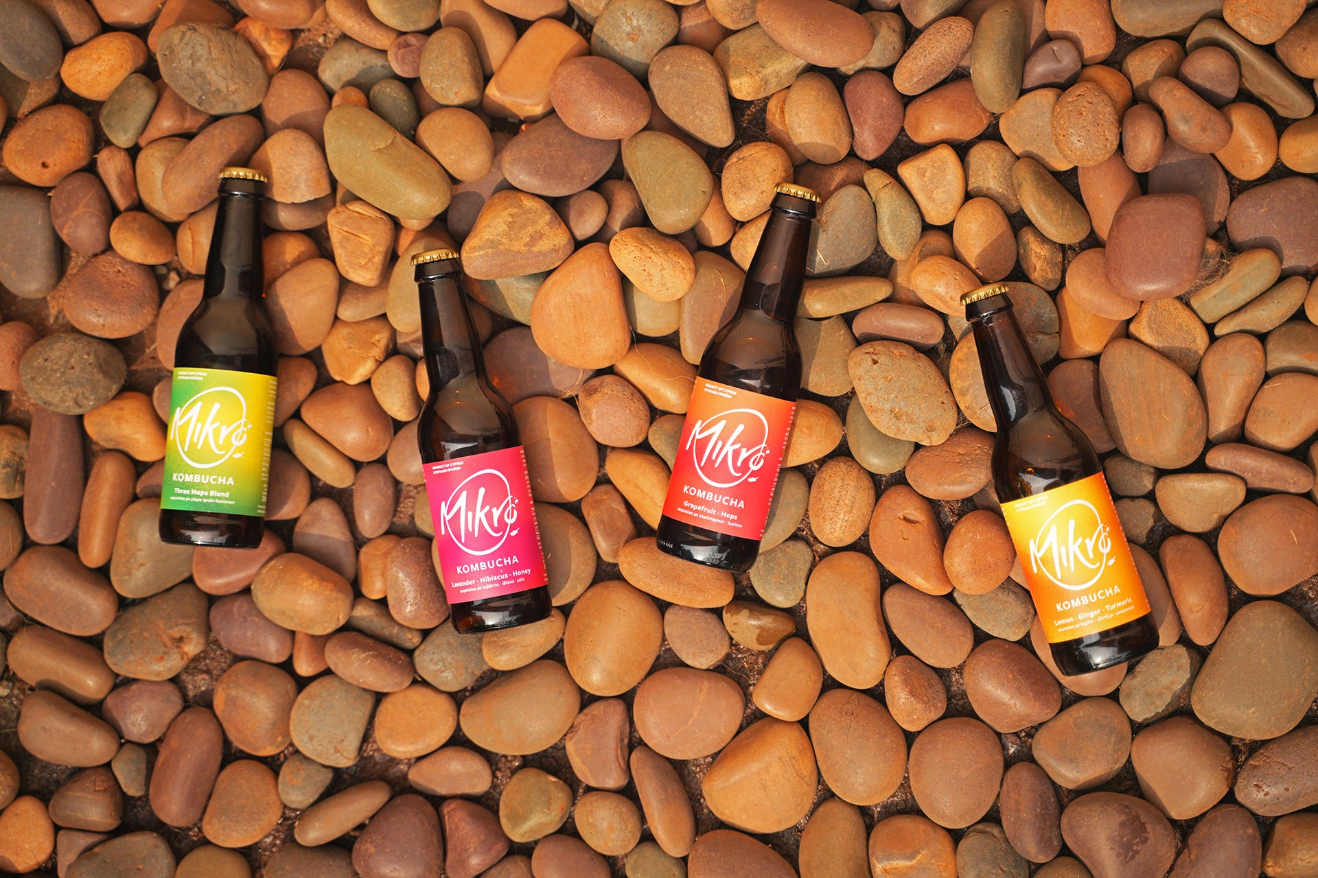 Four bottles of Mikro kombucha with colourful labels on a bed of multicolored stones.
